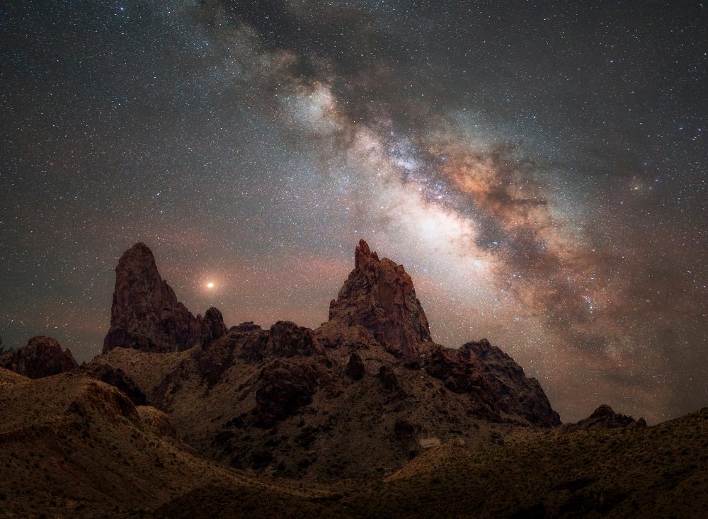 A stunning photo of the Milky Way galaxy over a desert landscape, with two rock formations resembling mule ears in the foreground. The night sky is filled with stars and nebulae, creating a contrast with the dark silhouette of the rocks and plants. The photo showcases the beauty and mystery of the cosmos, as well as the skill and creativity of Andy Morgan Photography.