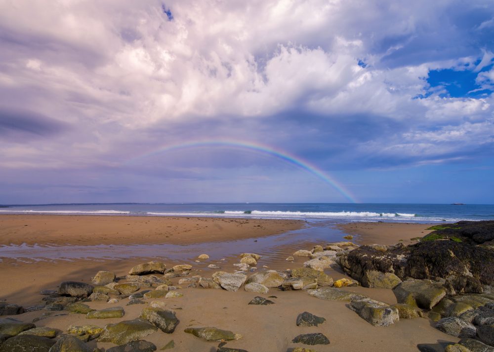 A full rainbow stretches across the horizon off a rocky beach on the coast of maine at Reid state park